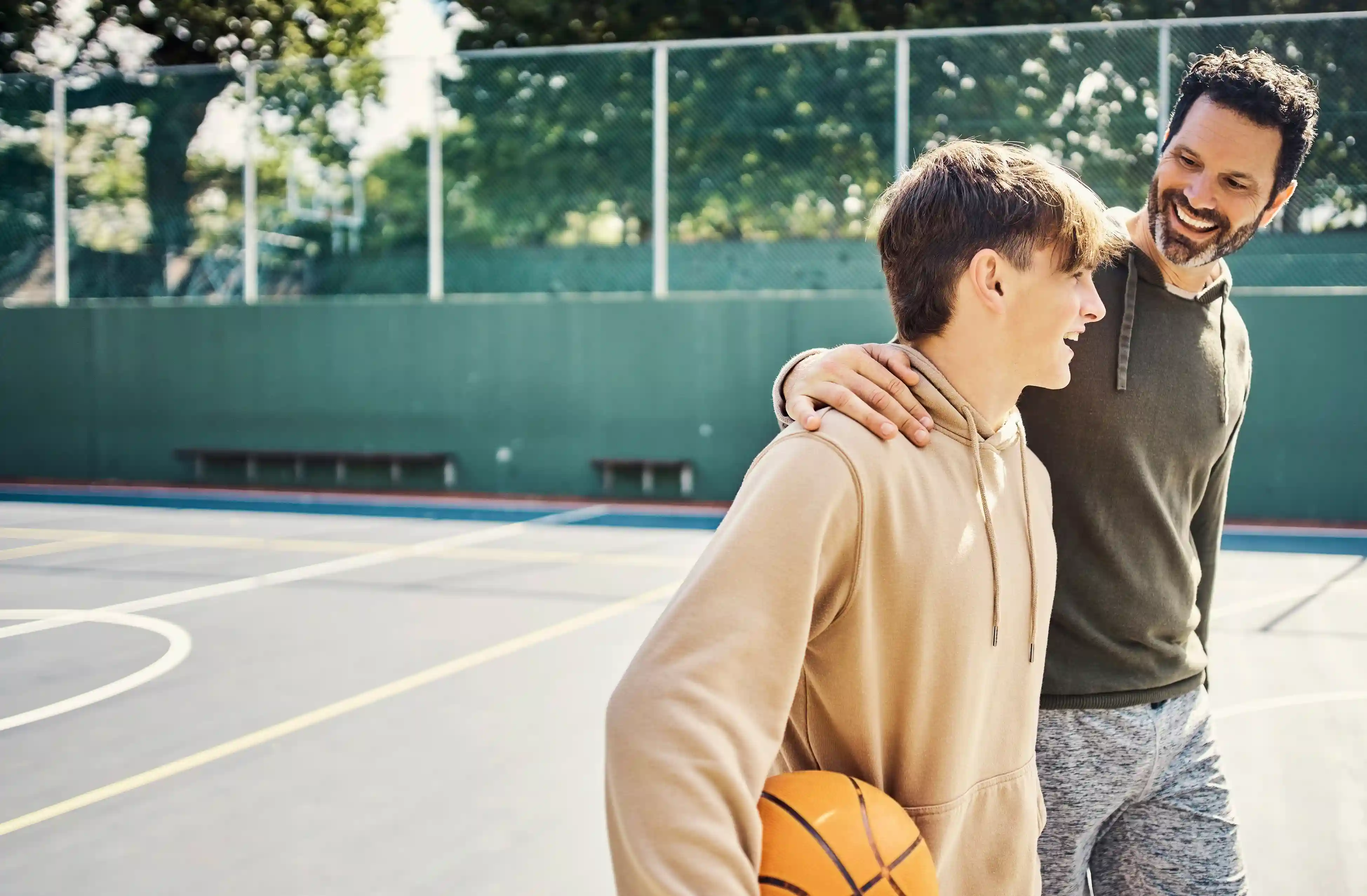sport- en bewegen als jongerenwerk van gro-up Een jongen loopt op een basketbalveld met een basketbal onder zijn arm en een jongerenwerker die zijn arm om zijn schouder heeft geslagen