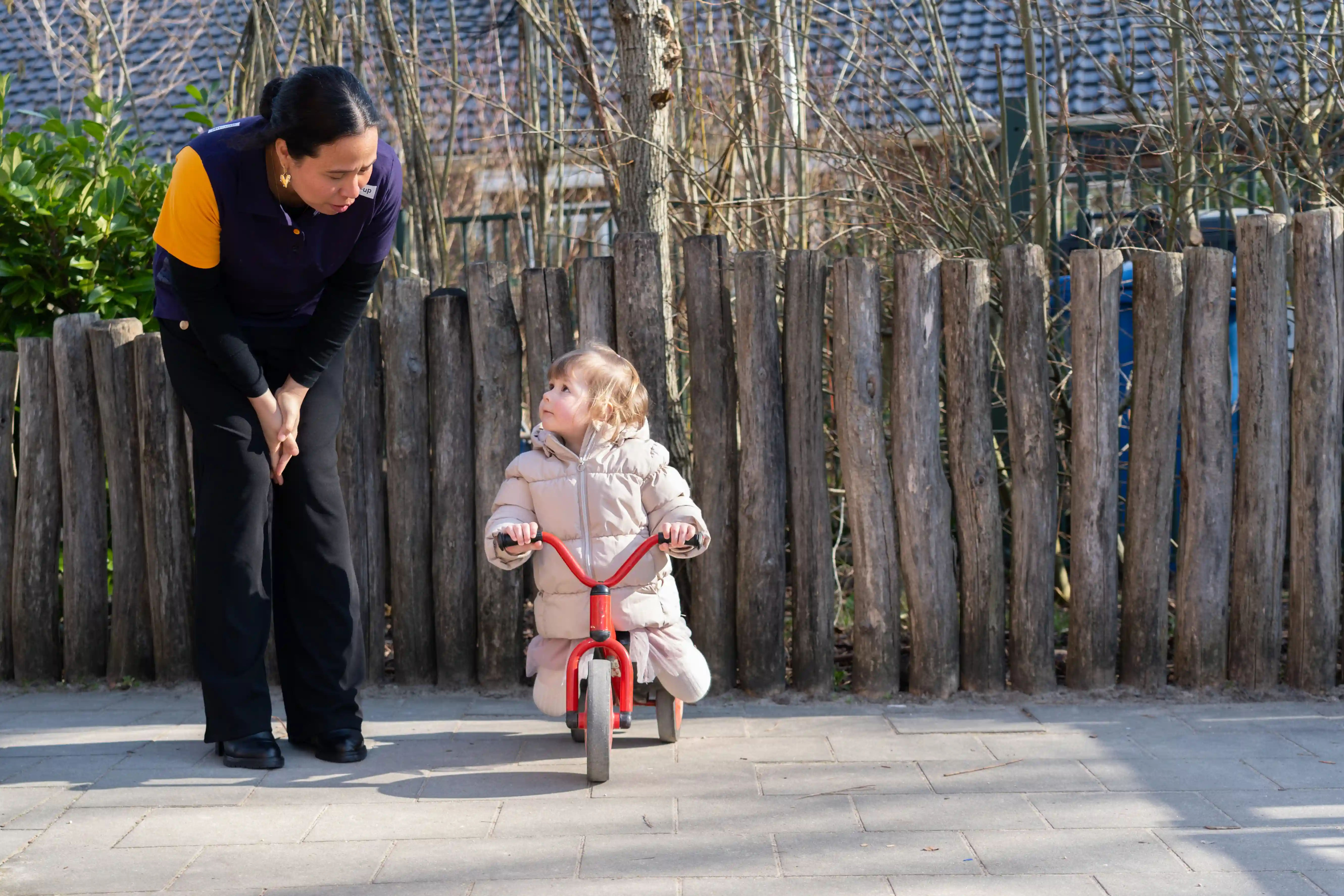Open Dag Peuteropvang Rotterdam Een pedagogisch medewerker helpt een peuter meisje die wil fietsen op een driewieler buiten bij een peuteropvang van grro-up