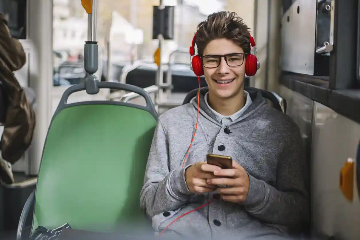Wat Is Online Jongerenwerk? Tienerjongen lacht in de bus lacht in de camera. Hij heeft rode koptelefoon op zijn hoofd en een smartphone in zijn handen.
